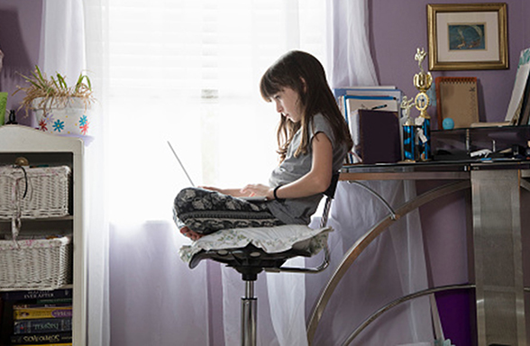 A young girl sits on a chair in a bedroom using a laptop on her lap