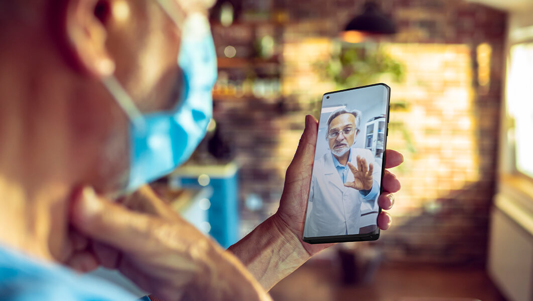 A senior wearing a facemask holds a smartphone during a telehealth visit with a doctor